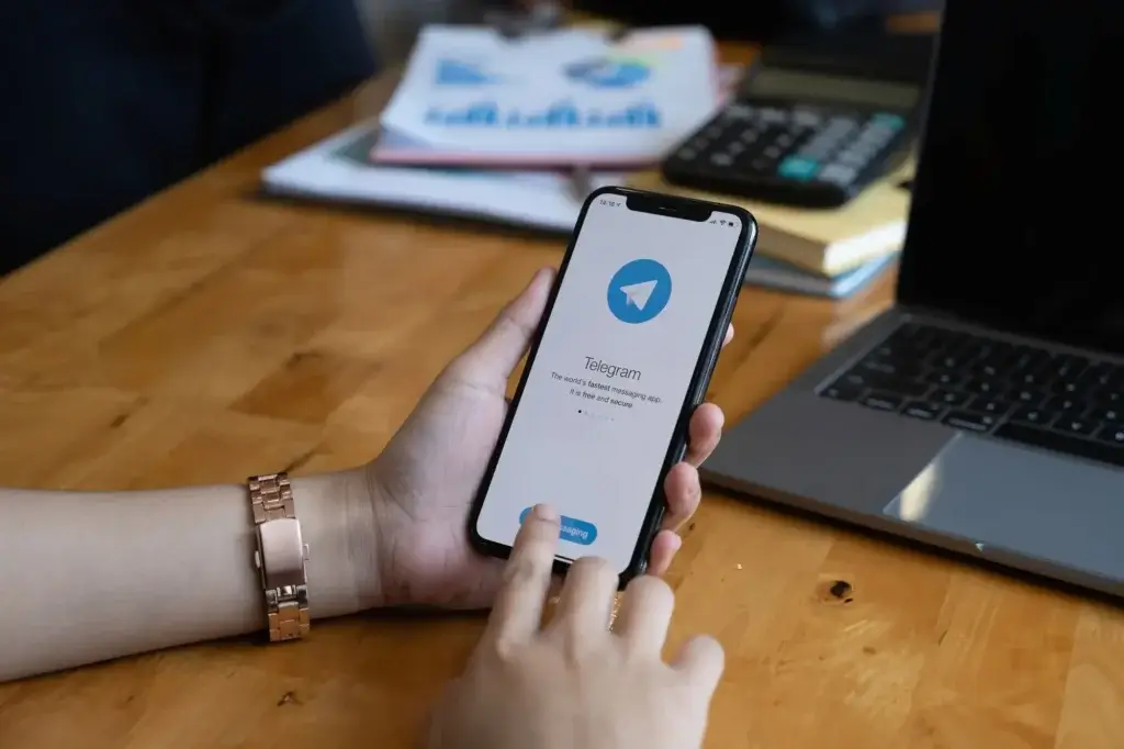 Person sitzt am Tisch, im Hintergrund ein Laptop und installiert die Messenger App Telegram am Handy.