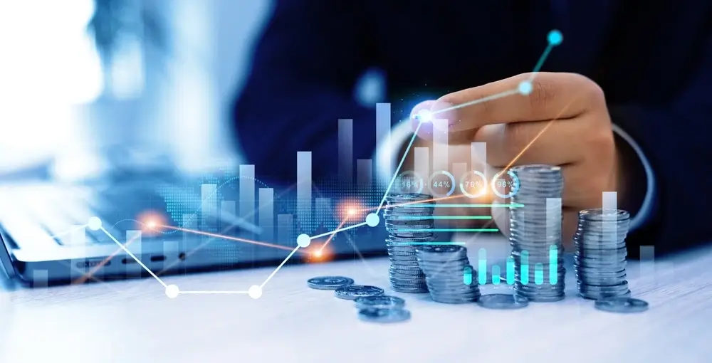 Ein Mann stapelt Geldmuenzen waehrend er am Laptop sitzt und seine Finanzen analysiert, deren Entwicklung als Graph dargestellt wird.